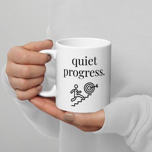 Könnte beinhalten: Weiße Keramik-Tasse mit dem schwarzen Schriftzug "quiet progress". Darunter befindet sich eine schwarze Grafik einer Person, die eine Treppe zu einem Ziel hinaufsteigt. Die Tasse wird von einer Person gehalten.
