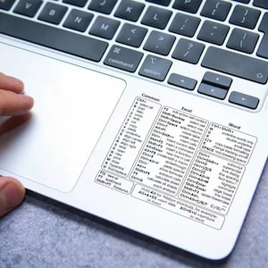 Könnte beinhalten: Ein weißer Laptop mit schwarzer Tastatur und einem Aufkleber mit Tastenkombinationen. Der Aufkleber enthält Verknüpfungen für gängige Funktionen, Excel und Word. Eine Hand ist sichtbar.