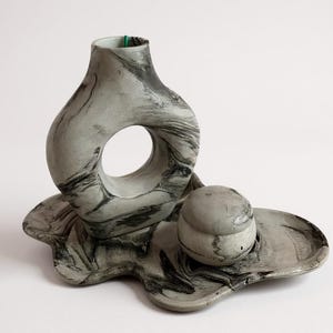 May include: A grey and black marbled ceramic decorative set. The set includes a vase with a large circular opening, a small round container with a lid, and a uniquely shaped tray. The vase is approximately 18 cm tall.