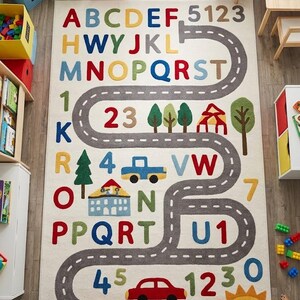 May include: A rectangular area rug featuring a cream background with a winding grey road, colourful alphabet letters, numbers, and various icons including trees, buildings, a blue car, a red car, and a yellow sun printed across the surface.