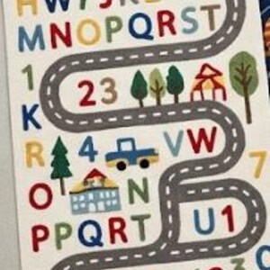 May include: A rectangular educational wall decal featuring a winding grey road with white dashed lines. The design includes colourful letters of the alphabet, numbers, green trees, a blue truck, a house, a red car, and a yellow sun in the corner.