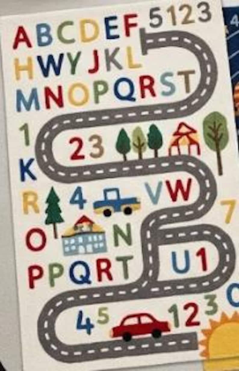 May include: A rectangular educational wall decal featuring a winding grey road with white dashed lines. The design includes colourful letters of the alphabet, numbers, green trees, a blue truck, a house, a red car, and a yellow sun in the corner.