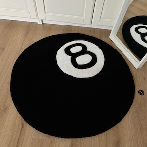 May include: A round black rug with a white number eight in the center, resembling an eight ball. The rug is on a wooden floor, and a mirror reflects the rug's design. The rug is a decorative item.