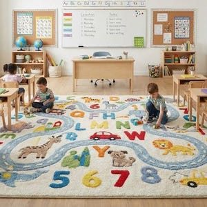 May include: A classroom scene featuring a large, colorful rug with a road design, animals, numbers, and letters. Wooden desks and chairs are arranged around the rug. The room includes a whiteboard, bulletin boards, and educational materials.