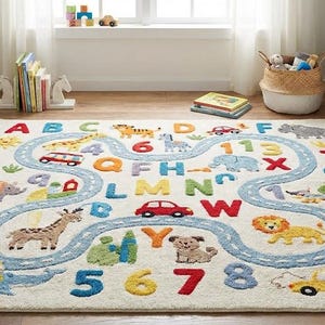 May include: A colorful children's rug with a road design, featuring letters, numbers, and animal illustrations. The rug has a cream background with a blue road winding through the center. Various vehicles and animals are depicted along the road.