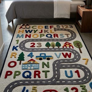 May include: A rectangular area rug with a cream-coloured background featuring a winding grey road, colourful raised letters of the alphabet, numbers, and various icons including trees, houses, and cars in red, blue, green, and yellow hues.