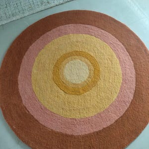 May include: A round rug with concentric circles in shades of brown, pink, yellow, and cream. The rug has a textured surface and a retro design. The rug is placed on a light blue surface. Another rug is partially visible in the background.