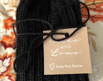 May include: A black knitted beanie with a ribbed texture, tied with a black yarn string holding a square tan paper tag. The tag features the white printed text "made with Love" and "Knitsy Bitsy Blankets" with a small white heart icon.