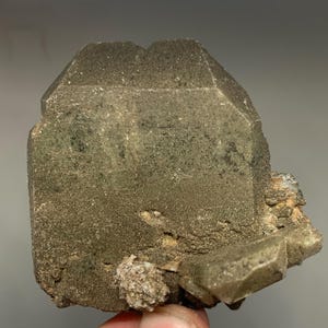 Puede incluir: Un espécimen mineral rugoso y texturizado con un tono gris verdoso oscuro y apagado. El cuerpo principal tiene una forma geométrica y bloqueada con una apariencia ligeramente desgastada. Se adjuntan formaciones minerales más pequeñas y claras.