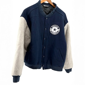 May include: A navy blue and grey varsity jacket featuring the Dallas Cowboys logo. The jacket has a button-up front, grey sleeves, and a ribbed collar and cuffs. The logo is a white star within a white circle with the words "DALLAS COWBOYS".