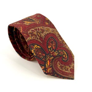Puede incluir: Una corbata con un patrón de cachemira detallado en tonos rojo intenso, marrón oscuro y amarillo dorado. La tela muestra intrincados motivos florales y de gota tejidos en todo el diseño, con la corbata enrollada en la parte superior.