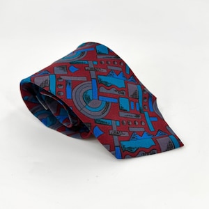 Könnte beinhalten: Eine Krawatte mit einer tiefroten Grundfarbe und einem sich wiederholenden geometrischen Muster in leuchtenden Blau-, Hellblau- und Grautönen. Das Design umfasst verschiedene Formen wie Kreise, Rechtecke und gezackte Linien auf dem Stoff.