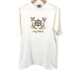 Puede incluir: Una camiseta blanca de manga corta con un gráfico central de dos anclas doradas cruzadas detrás de una rosa de los vientos con las letras N, E, W y S. Debajo del gráfico, el texto Key West está impreso en una fuente cursiva dorada.