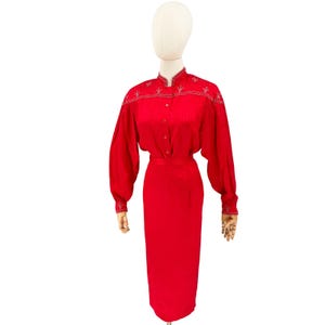 Puede incluir: Un vestido rojo vibrante de manga larga con cuello alto, cierre frontal de botones y bordados plateados detallados en los hombros y puños. La prenda presenta una cintura entallada y una falda recta que llega hasta la altura de la rodilla.