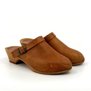May include: A pair of brown leather clogs with wooden soles. The clogs have a buckle closure and a small heel. The shoes are a light brown colour and appear to be vintage.
