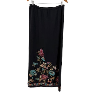 Pode incluir: Uma saia comprida preta com um padrão floral bordado na parte inferior. O bordado inclui flores e folhas em tons de rosa, turquesa e dourado, com uma borda horizontal decorativa que percorre toda a bainha inferior da peça.