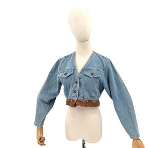 May include: A light blue denim cropped jacket featuring long sleeves, a V-neckline, and a front button closure. The jacket includes two chest pockets with flaps and a brown lace trim detail along the bottom hemline of the garment.