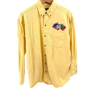 May include: A long-sleeve button-down shirt in a solid light yellow color. The shirt features a button-down collar and a single chest pocket. An embroidered patch on the pocket displays the word READING with book icons and the text POETRY, FICTION.