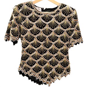 Puede incluir: Top negro y dorado con lentejuelas y un diseño festoneado. La parte superior de manga corta presenta un cuello redondo y un cierre de cremallera en la espalda. El diseño incluye un patrón repetitivo en forma de abanico con lentejuelas doradas y blancas.