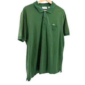 May include: A solid green short-sleeve polo shirt featuring a classic collar, a three-button placket, and a small chest pocket. The iconic green crocodile logo is embroidered on the pocket, and the interior label displays the Lacoste brand name.
