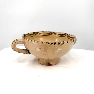 May include: A beige ceramic cup with a handle and a pedestal base. The rim is decorated with a scalloped pattern in dark brown and black. The handle and rim have black and brown accents. The cup has a rustic, handmade appearance.