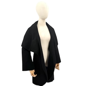 May include: A solid black open-front coat featuring a wide, draped shawl collar and long, straight sleeves. The garment has a smooth, uniform texture and a structured silhouette that falls to the mid-thigh, designed without buttons or fasteners.