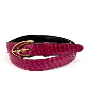 Puede incluir: Un cinturón de color magenta vibrante con un patrón texturizado similar a escamas en la correa. El cinturón se asegura con una hebilla de metal dorado pulido en forma de ancla o flecha estilizada e incluye una trabilla de cuero a juego.