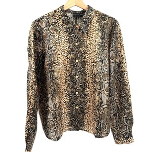 Puede incluir: Camisa abotonada de manga larga con estampado de piel de serpiente en tonos marrón, beige y negro. La camisa está hecha de un material ligero y cuelga de una percha de madera.