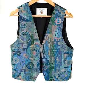 Pode incluir: Um colete sem mangas com um padrão vibrante estilo tapeçaria em tons de azul, verde e rosa. O tecido apresenta formas geométricas e motivos circulares. Uma etiqueta branca no decote interior exibe o texto ANTER FASHION.
