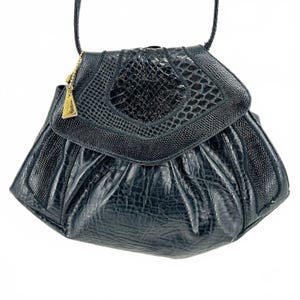 May include: A black handbag featuring a textured, reptilian-patterned flap and a gathered, crinkled body. The bag includes a thin shoulder strap and a small, triangular gold-toned metal charm attached to the side by a delicate ball chain.