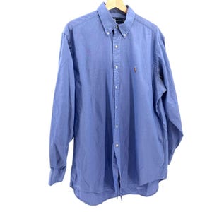 以下が含まれることがあります： 木製のハンガーに掛けられた、長袖のライトブルーのボタンダウンシャツ。胸には小さな刺繍ロゴがあります。襟はボタンダウンです。シャツは柔らかく軽い生地で作られています。