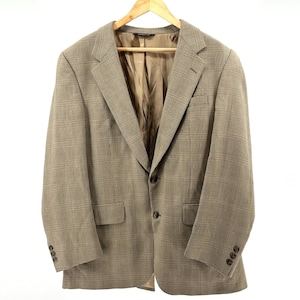 May include: A tan and brown plaid blazer with a two-button closure and a notched lapel. The blazer has two front pockets and buttons on the sleeves. It is hanging on a wooden hanger against a white background.