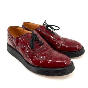 May include: A pair of burgundy patent leather oxford shoes featuring classic brogue detailing, perforated patterns, and black laces. The shoes are set on a thick, textured black rubber platform sole with a rounded toe and tan interior lining.