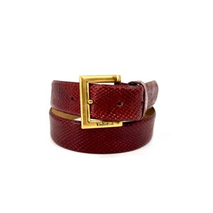 Puede incluir: Un cinturón de color rojo oscuro con una superficie texturizada y una hebilla rectangular de tono dorado. La hebilla presenta la palabra Unlisted grabada en el borde inferior. El cinturón está enrollado, mostrando la correa y los herrajes.