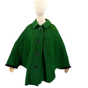 May include: A forest green wool-textured cape featuring a classic fold-over collar and four large, dark circular buttons down the front. The garment has a wide, flowing silhouette with dark trim along the sleeve openings, providing a structured look.