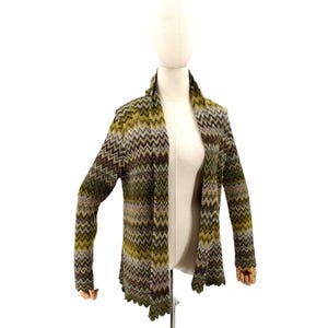 May include: A long-sleeved open-front cardigan featuring a multi-colored chevron pattern in shades of olive green, brown, tan, and cream. The garment has a shawl collar and a scalloped hemline along the bottom edge and the sleeve cuffs.