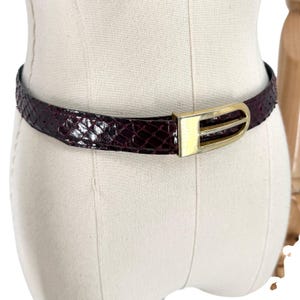 May include: A dark burgundy belt with a textured snakeskin pattern and a polished gold-tone metal buckle. The belt features a slim profile and a smooth, shiny finish, secured with a rounded, open-frame buckle design that sits centered on the strap.