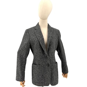 Könnte beinhalten: Ein grauer Tweed-Blazer mit Zwei-Knopf-Verschluss, zwei Vordertaschen und einem fallenden Revers. Die Jacke wird auf einer Schaufensterpuppe präsentiert. Der Stoff hat eine gesprenkelte Textur.