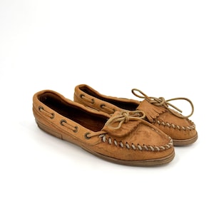 May include: A pair of tan suede moccasins featuring a whipstitch detail along the toe, a decorative fringe panel, and a lace-up closure with metal eyelets. The shoes have a low-profile design with a flat sole and a soft, textured exterior finish.