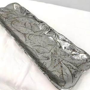 Puede incluir: Una bandeja rectangular de metal plateado con un diseño de peces en relieve. La bandeja tiene una forma ligeramente irregular y una superficie texturizada, con el patrón de peces que cubre toda la superficie.