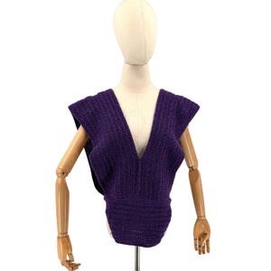 May include: A deep purple, sleeveless top with a plunging V-neck. The knitted garment has a fitted waist and broad shoulders, displayed on a mannequin. The top is made of a textured, possibly wool, material.