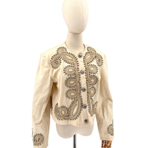 May include: Cream-colored jacket with ornate silver and gold embellishments. The jacket features intricate scrollwork and stud details around the collar, front, and sleeves. It has a round neckline and silver buttons. A unique, vintage fashion piece.
