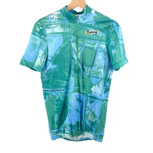 May include: A short-sleeve cycling jersey featuring a green and blue abstract geometric pattern. The shirt has a high collar with a central green zipper and a small white logo patch on the chest with the text Giessegi printed in black letters.