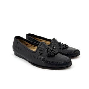 Puede incluir: Un par de mocasines de cuero negro con puntera cosida estilo mocasín, una borla decorativa en el empeine y un pequeño detalle de hebilla metálica. Los zapatos tienen un tacón bajo y un forro interior de color beige.