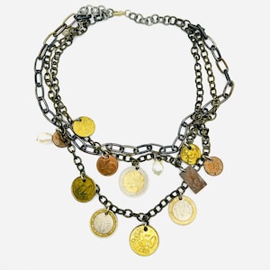 May include: A multi-strand necklace featuring dark metal chains adorned with various Euro coins, including 10, 20, and 50 cent pieces, alongside small white pearls. The design incorporates mixed metallic tones and a lobster claw clasp closure.