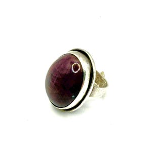 May include: A silver ring featuring a large, oval-shaped, deep burgundy cabochon stone set in a smooth metal bezel. The band of the ring has a textured, hammered finish, providing a contrast to the polished surface of the dark red gemstone.
