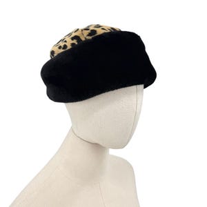 May include: A hat featuring a wide, solid black plush band around the base and a tan crown with a black leopard print pattern. The hat is constructed from a soft, textured material and sits on a neutral-colored mannequin head display.