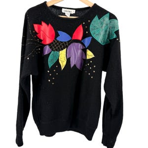 May include: A black long-sleeve sweater featuring a crew neckline and a label reading CRISTINA. The front is decorated with colorful leaf-shaped appliqués in red, blue, purple, yellow, and green, accented by small gold-toned studs and stitching.