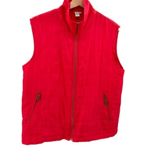 May include: A bright red quilted vest featuring a full-length front zipper closure, a high stand-up collar, and two side pockets with zipper closures. The garment has a grid-like stitched pattern across the front and a white interior label.