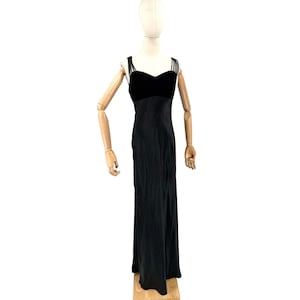 Puede incluir: Un vestido negro largo hasta el suelo con escote corazón, cuerpo de terciopelo y falda con acabado satinado. La prenda incluye múltiples tirantes finos en cada lado, creando un detalle tipo jaula que conecta el cuerpo con la espalda.